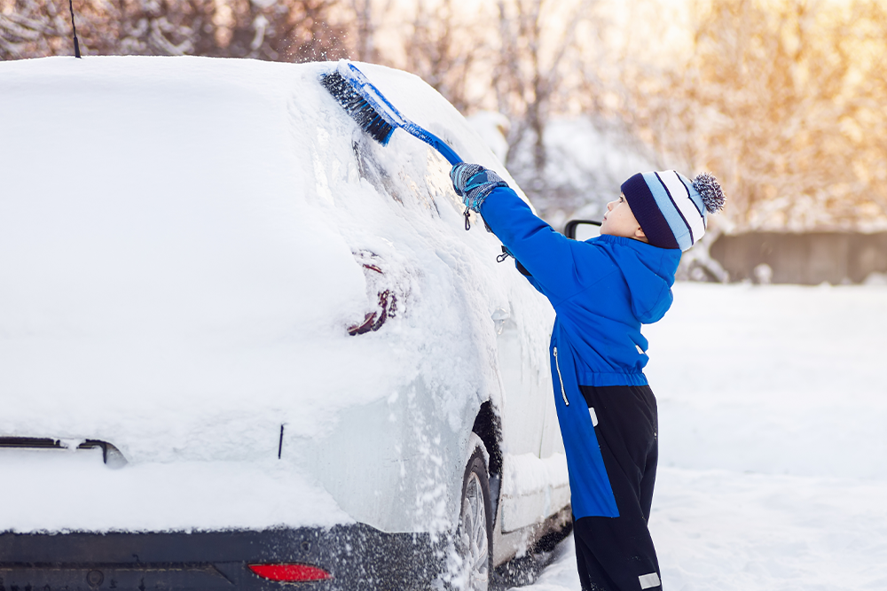 A young boy uses a snow brush to clear snow from a vehicle's rear window artboard-13.png