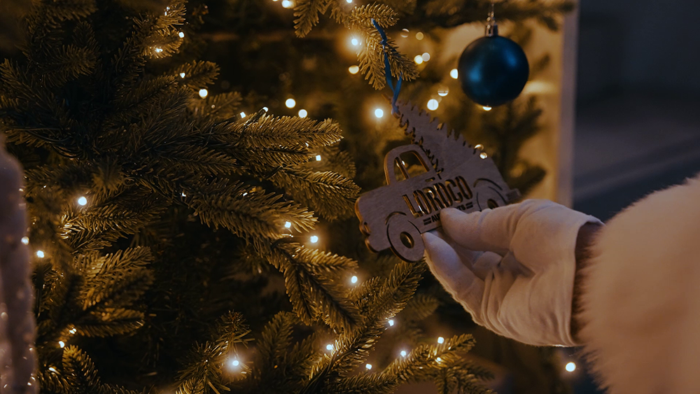Santa hangs an ornament shaped like a pickup truck carrying a tree in it's bed on a tree lit with warm, white lights