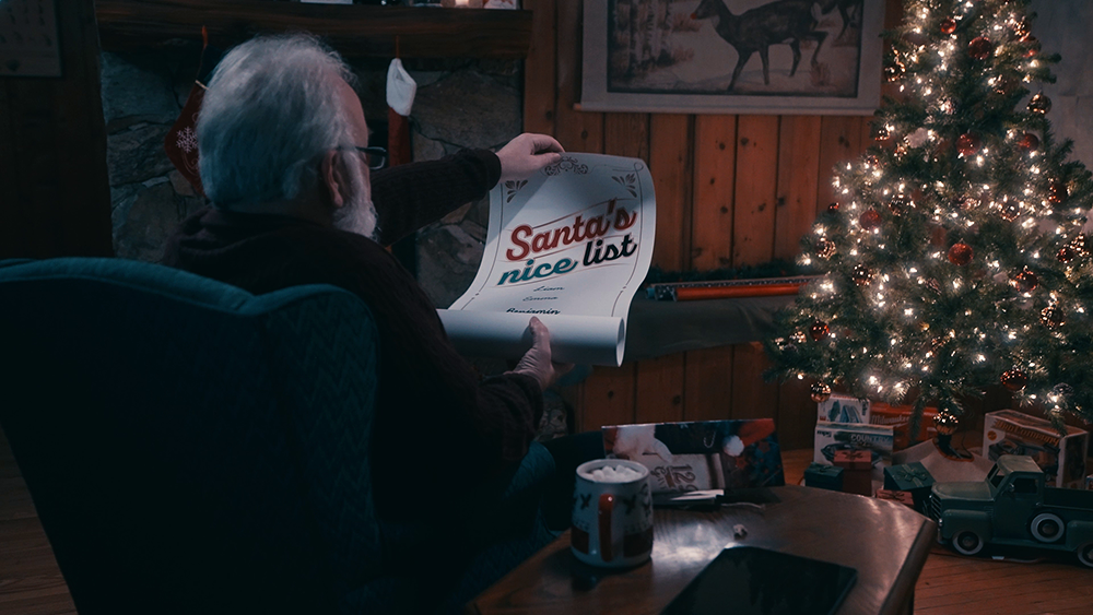 Santa looks over his "nice list" while sitting in a comfy chair facing a Christmas tree aglow with lights