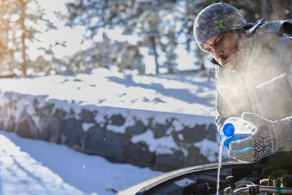 A man stands by a car with it's trunk open and pours washer fluid into the washer fluid reservoir 